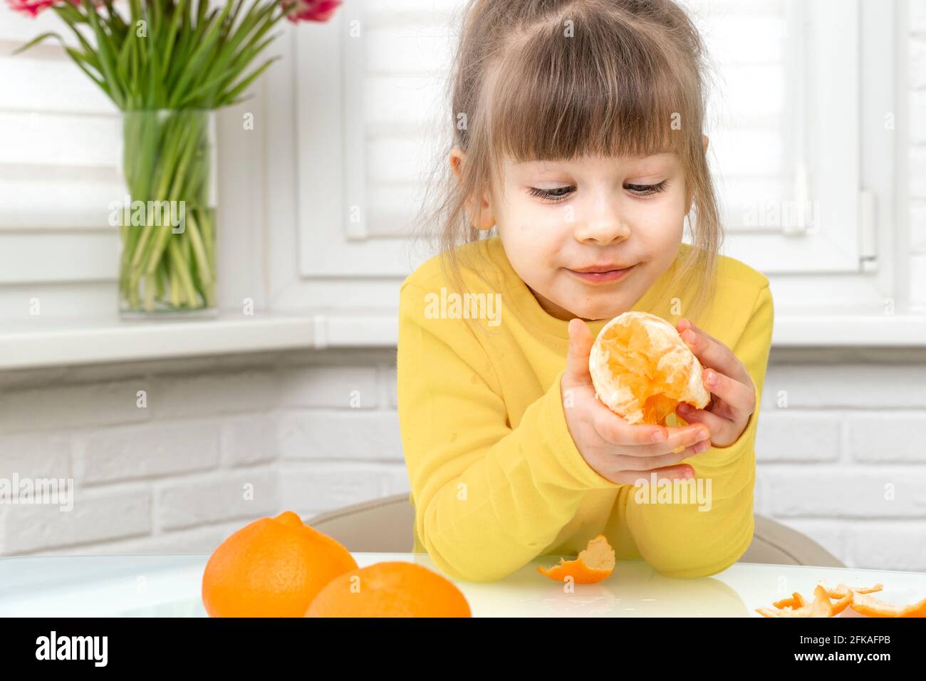 happy little girl eating big mandarin whole while sitting at table in white kitchen Stock Photo