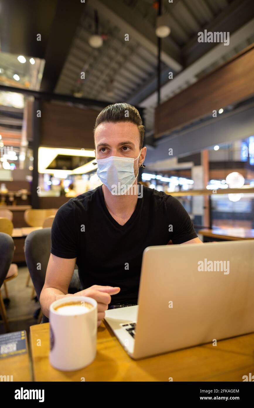 Man sitting at coffee shop wearing face mask and using laptop computer while thinking vertical shot Stock Photo