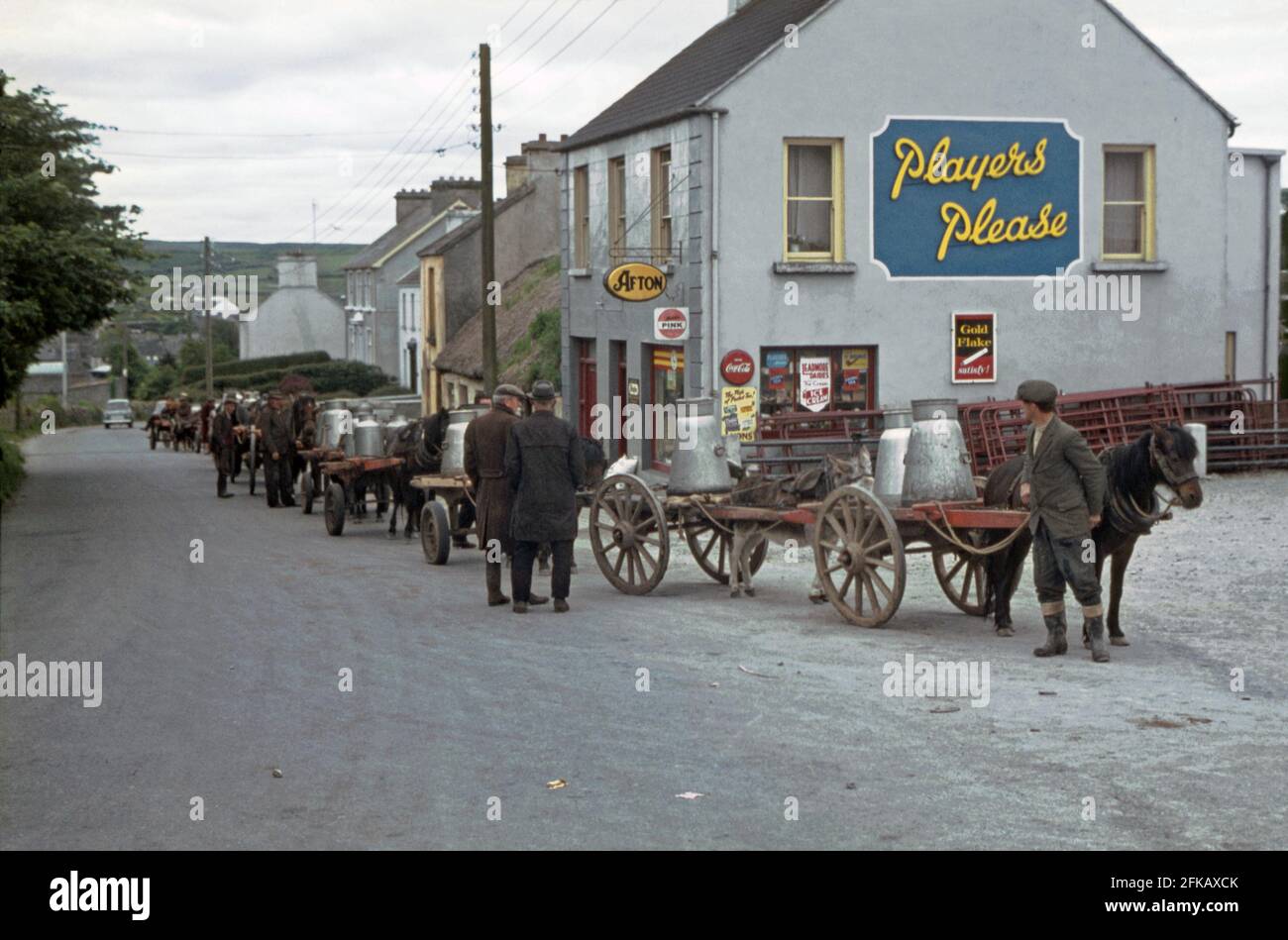A queue is forming outside the local dairy in Church Street on the outskirts of Ennistimon (Ennistymon or Inis Díomáin), County Clare, Ireland in the 1960s. The farmers have brought their milk for processing in churns by cart, pulled by horse or donkey. Donkeys were a common site on farms in Ireland. The store next to the dairy has a large Players cigarette advertising sign on the side wall. The site of the dairy is now a farm store. This image is from an old amateur colour transparency – a vintage 1960s photograph. Stock Photo