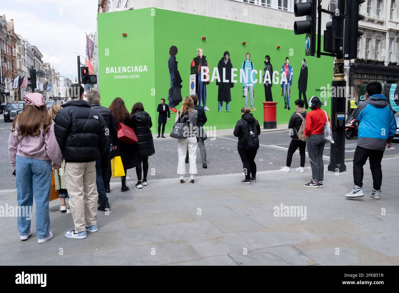 Shoppers cross the road towards the temporary hoarding for Balenciaga, a  retail space which is opening soon on Bond Street, on 27th April 2021, in  London, England. Balenciaga is a fashion house