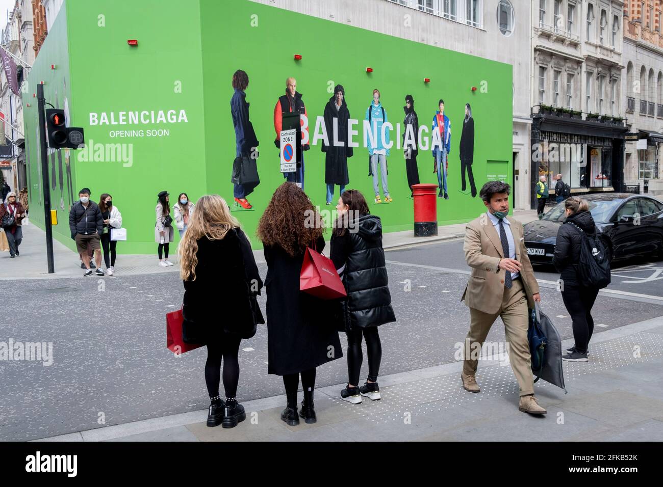 Shoppers cross the road towards the temporary hoarding for Balenciaga, a  retail space which is opening soon on Bond Street, on 27th April 2021, in  London, England. Balenciaga is a fashion house