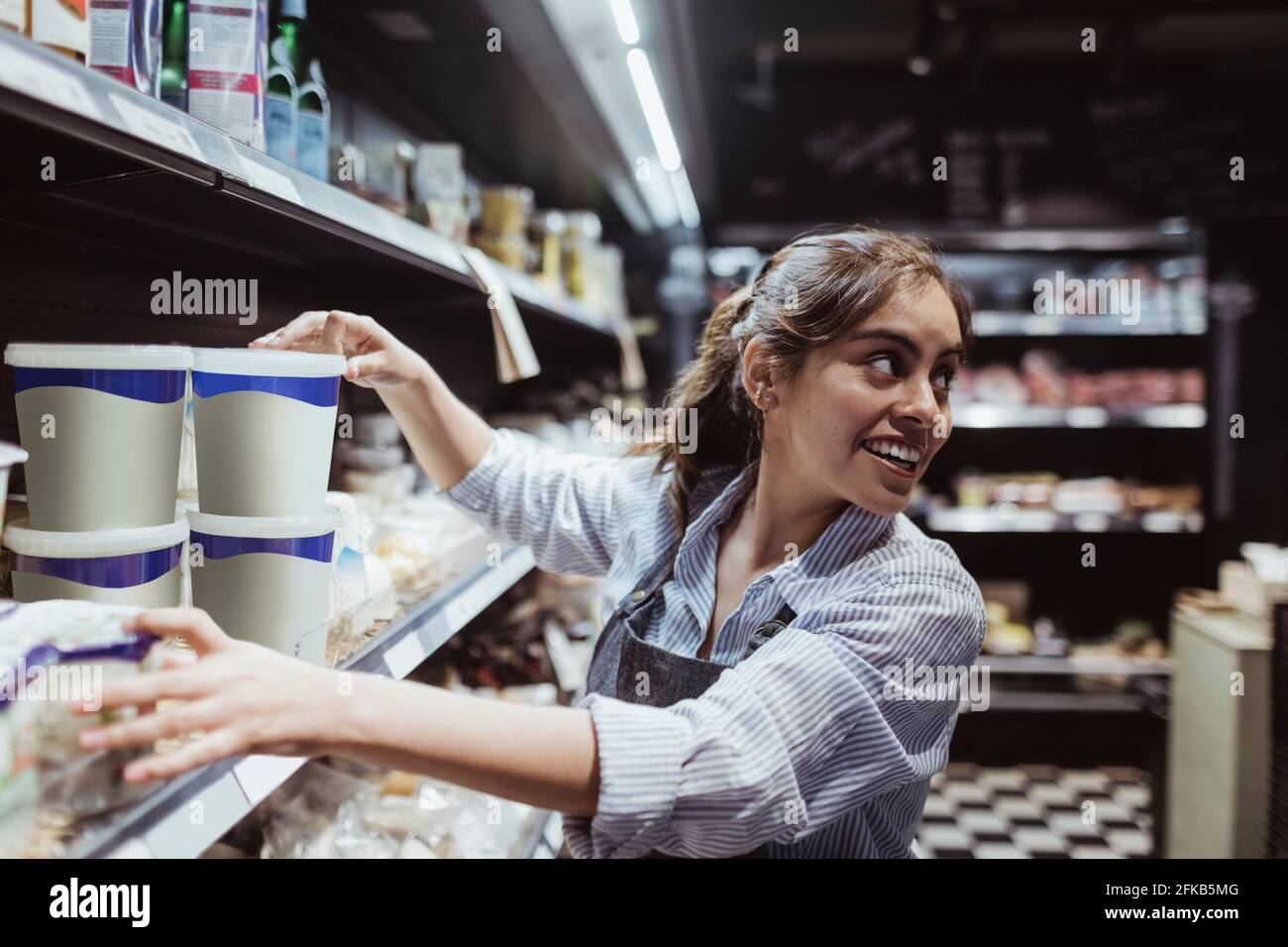 Young female owner looking over shoulder while arranging product on rack at delicatessen shop Stock Photo