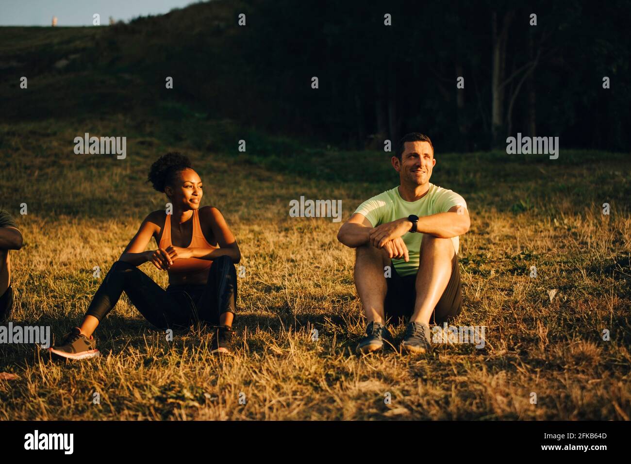 Male and female athletes looking away while sitting on grass during sunset Stock Photo