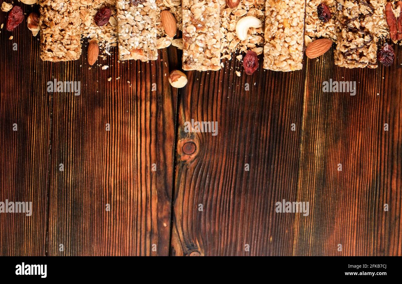Variety of granola energy bars in row with scattered mixed nuts, cereals, cranberry on brown wood textured table background. Healthy nutritious vegan Stock Photo