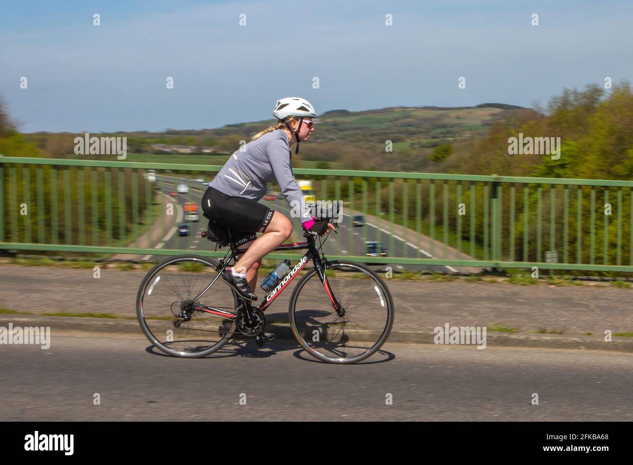 Female cyclist riding Cannondale sports road bike, blurred, low shutter speed, motion blur,  blurring, moving, panning moving road race bikes, cycling, carbon fibre bicycle frames, monocoque composite bike frame; fast cyclists racing at speed, bikers on countryside route crossing motorway bridge in rural Lancashire, UK Stock Photo