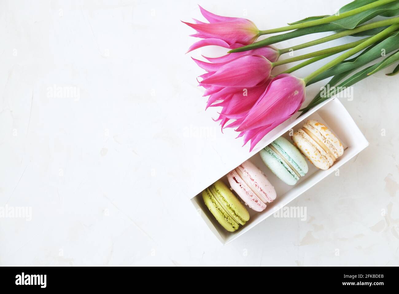 Beautiful feminine composition w/ box of French macaroon sweets pastel colors & purple tulip flowers blossoms on stucco background. 8 march women's da Stock Photo