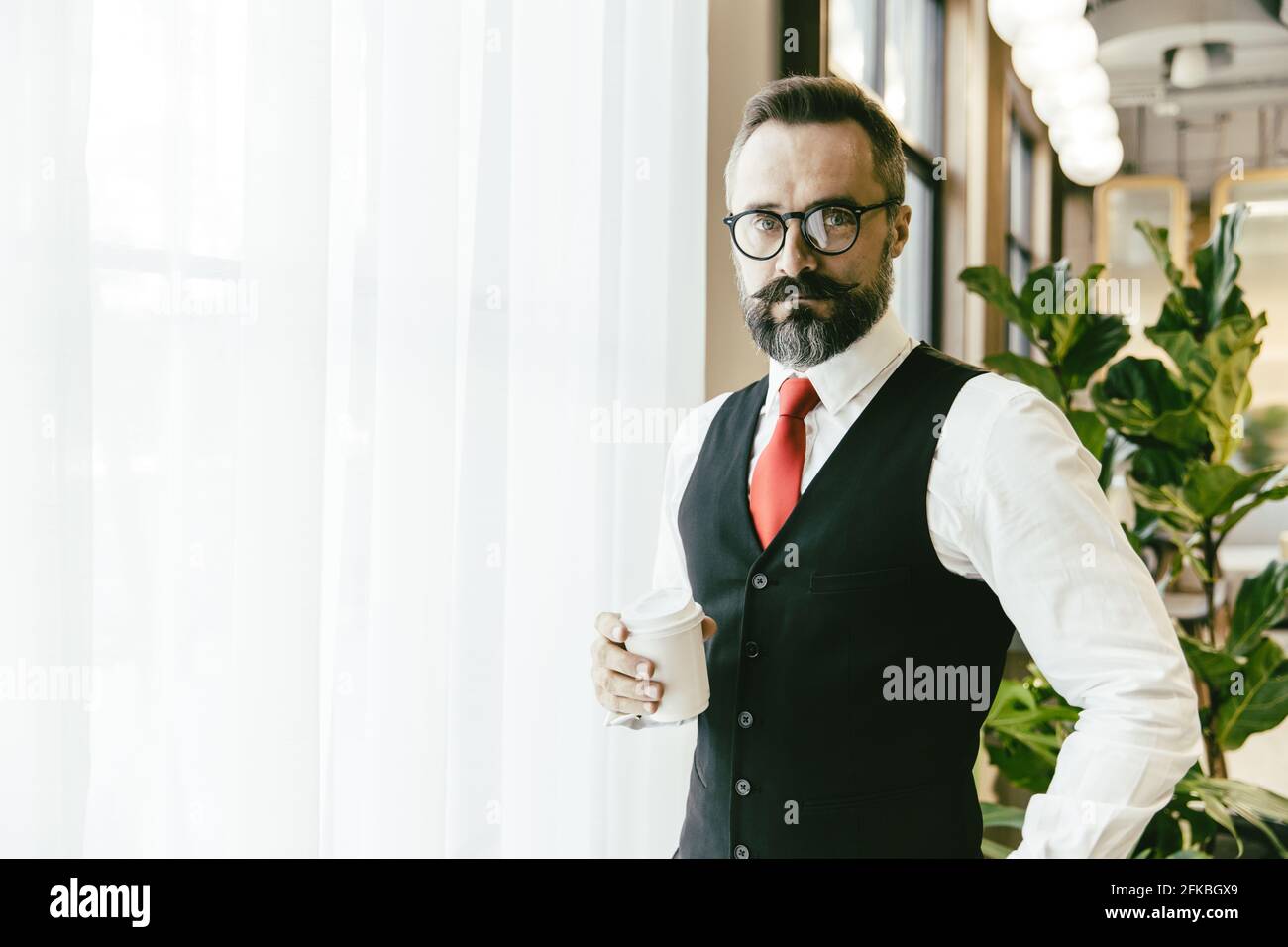 Portrait serious professional conservative adult model businessman Latin beard standing hand holding coffee cup in modern office with copy space for t Stock Photo