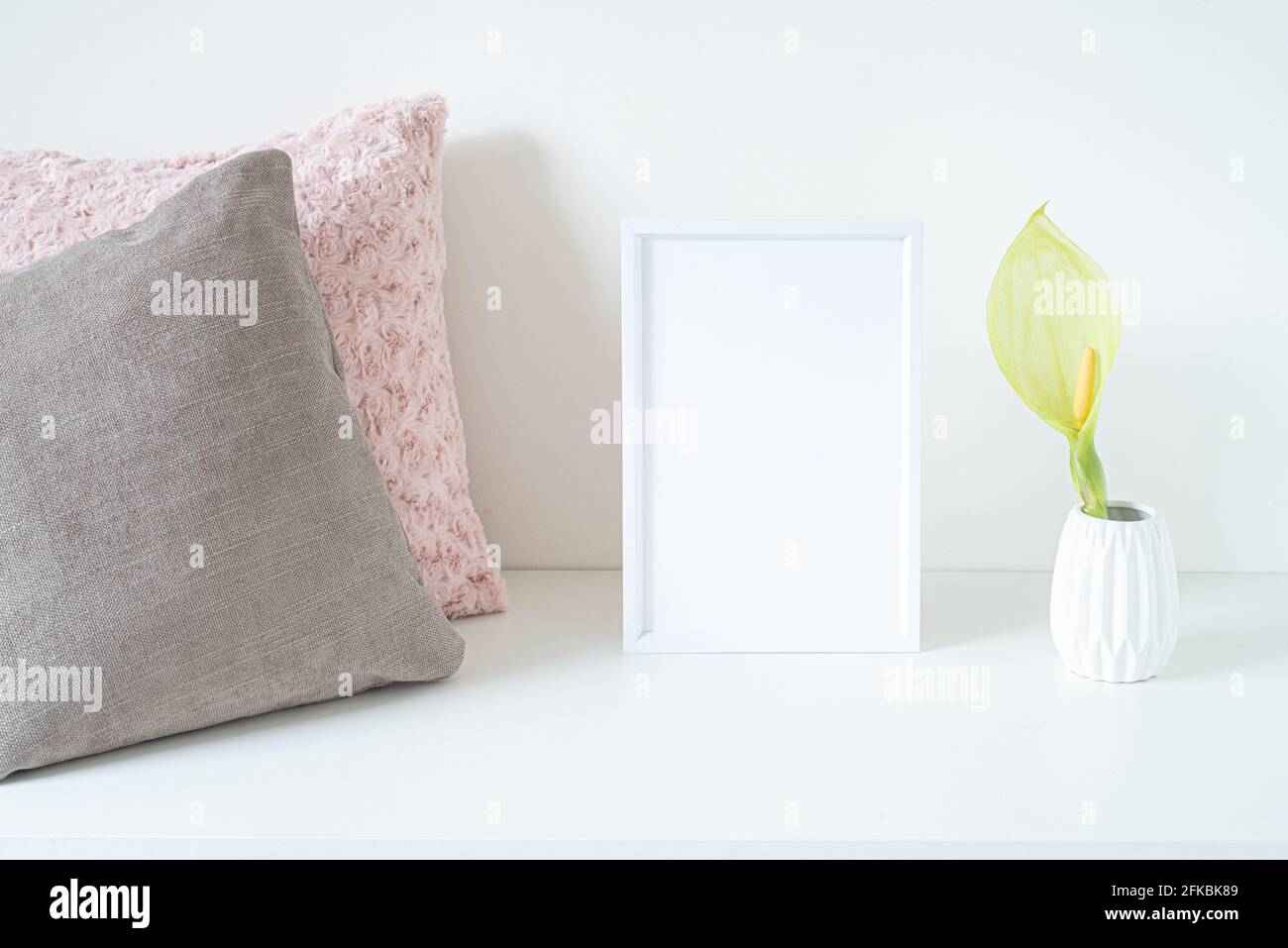 Empty white frame mockup on white table. Soft colored cushions and white vase with an arum italicum flower. Modern and minimal lifestyle. Scandinavian Stock Photo