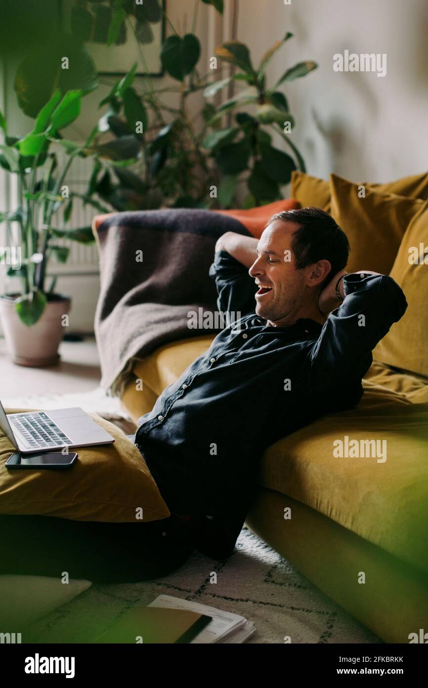 Male entrepreneur yawning with hands behind head in living room at home Stock Photo