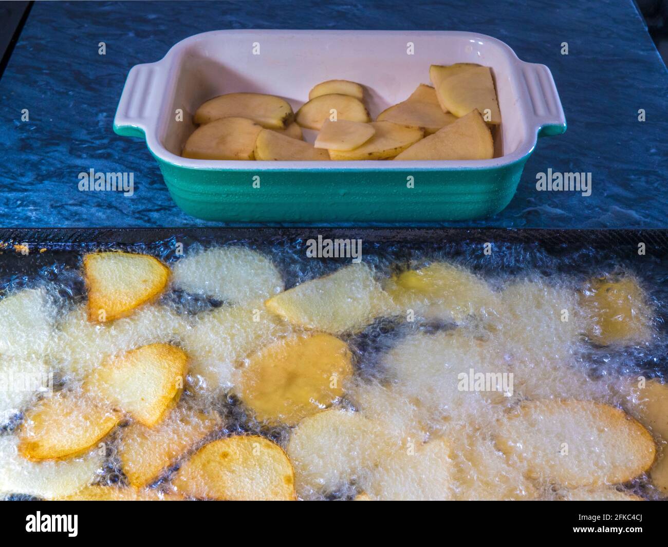 Closeup POV shot of thinly sliced pieces of raw potato in a dish, ready to cook, with some already frying in a flat pan of bubbling hot oil. Stock Photo