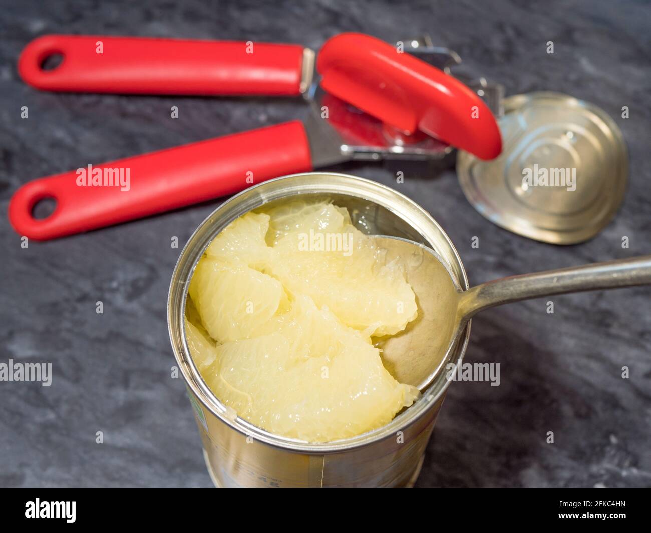 Closeup POV overhead shot of a large spoon lifting some grapefruit slices in juice from a newly opened tin can, with the opener and lid nearby. Stock Photo