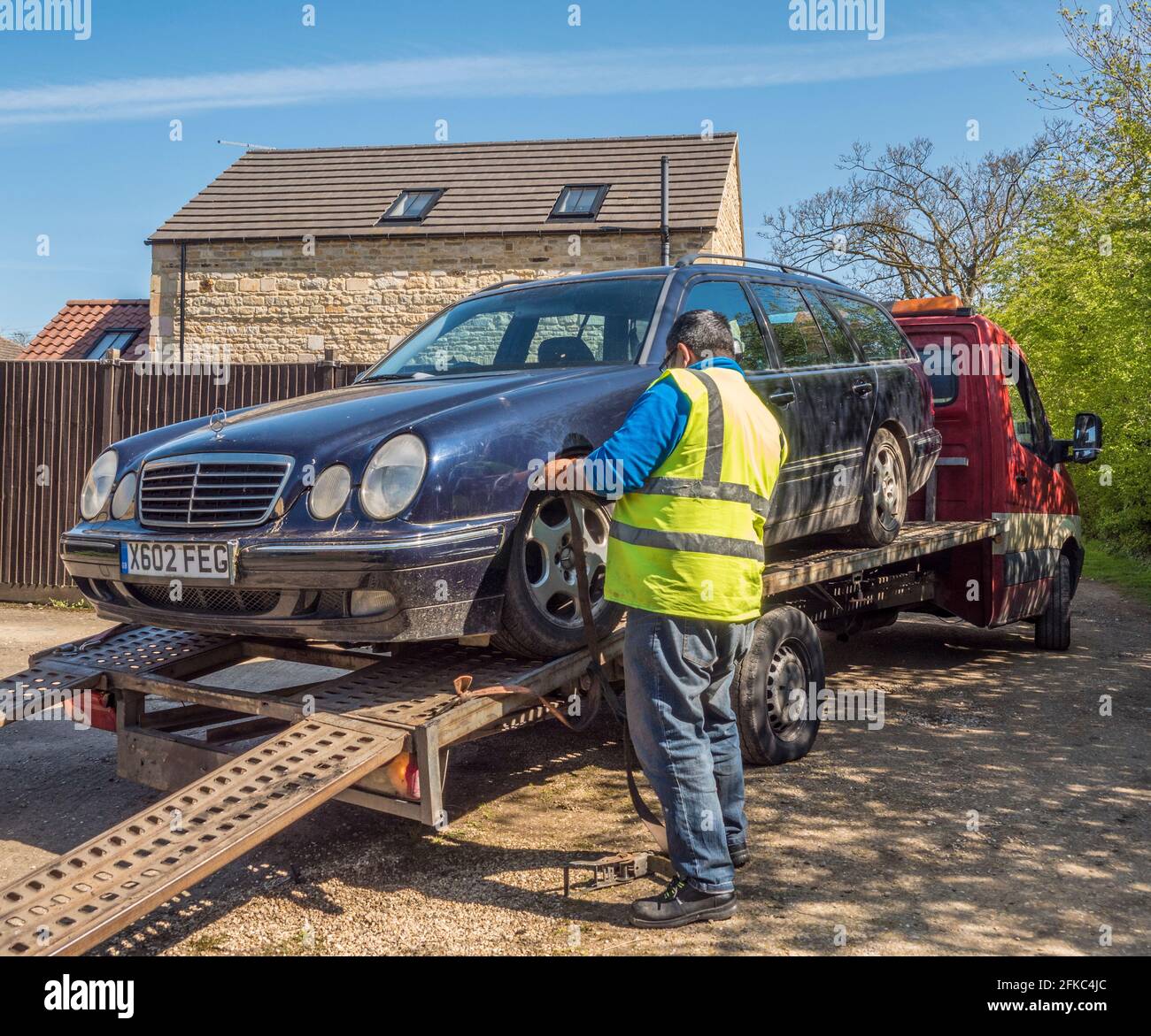An old Mercedes estate car on a flatbed truck / recovery vehicle, almost ready to be taken away, with the driver tightening a wheel securing strap. Stock Photo