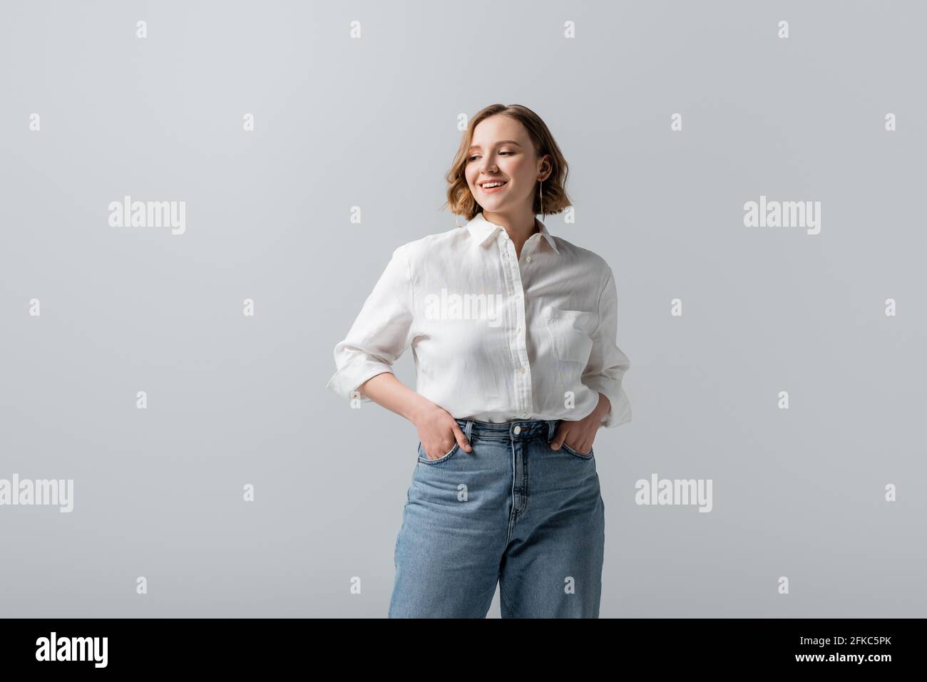 smiling overweight young woman in jeans posing with hands in pockets isolated on grey Stock Photo