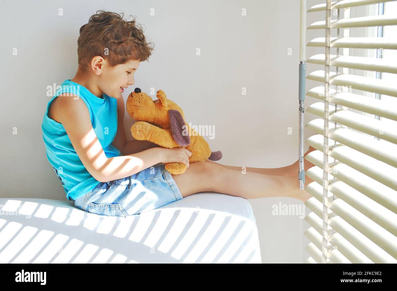 A cute little boy with curly hair sits on white sofa in sunglasses with his toy dog and smiles Stock Photo