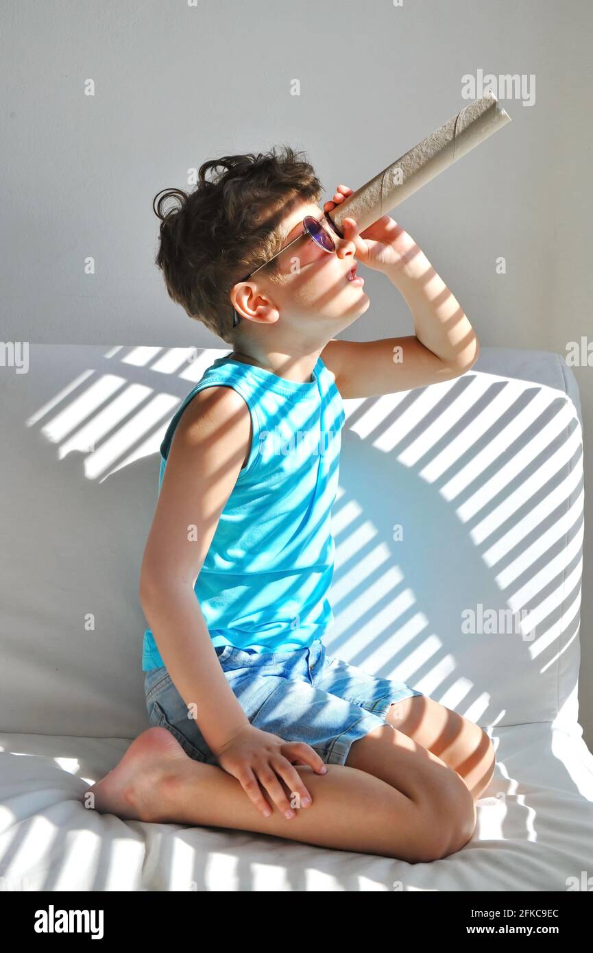 A cute little boy sits in a room and looks through a paper spyglass. Stock Photo