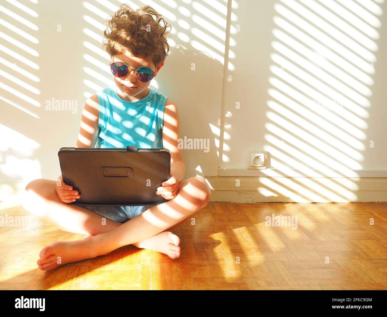 A cute little boy in blue sunglasses with a laptop is sitting against a white wall. On it and on the wall there are shadows from the blinds Stock Photo