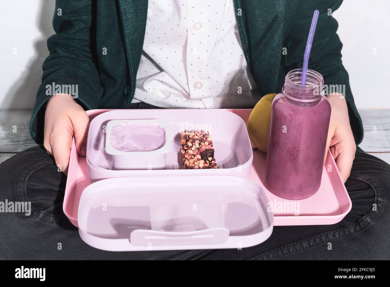 A schoolboy sits on the floor in a lotus position with a tray in his hands with a healthy snack, yogurt, a cereal bar, an apple and an oatmeal cocktai Stock Photo