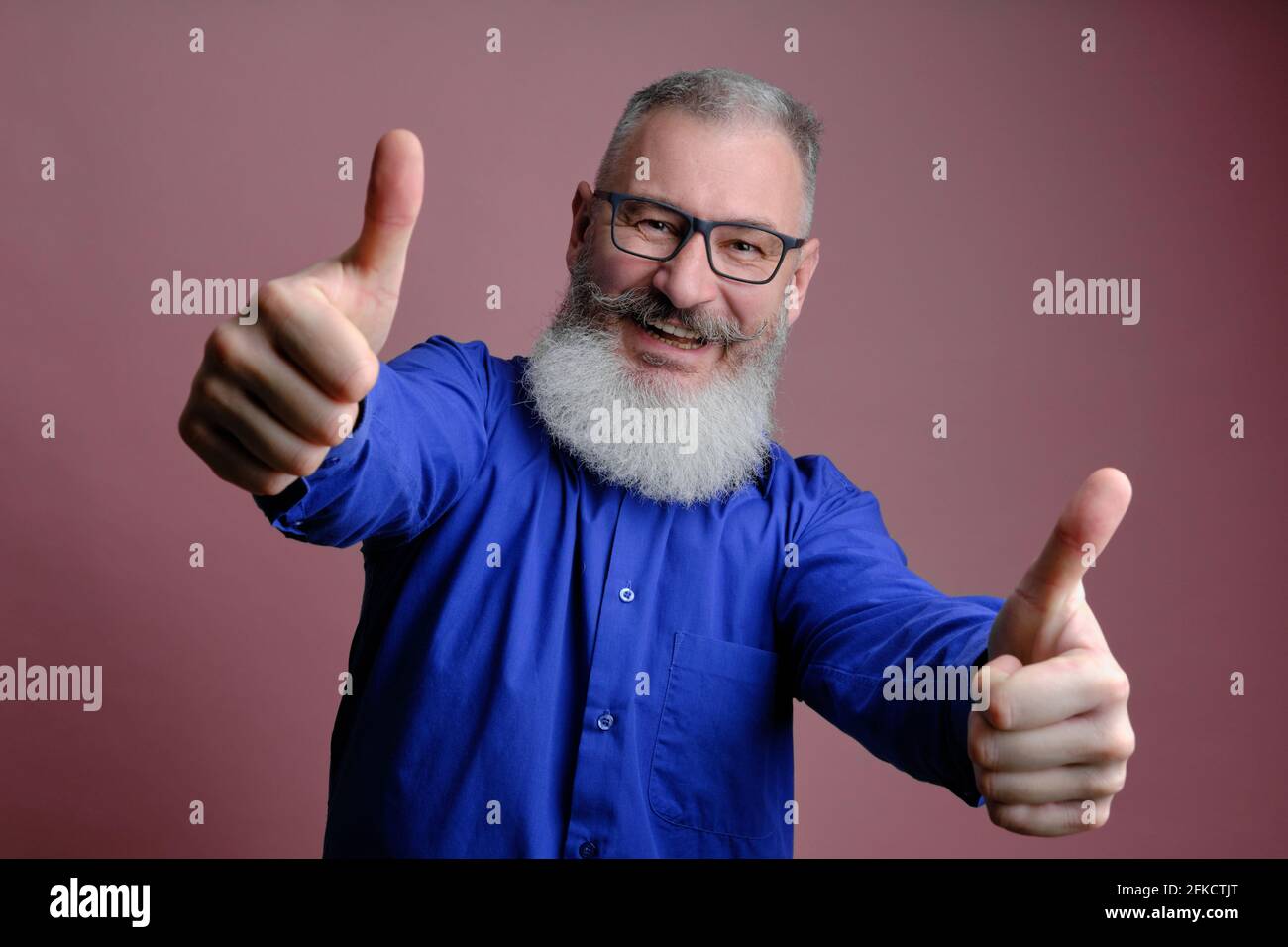 Portrait of mature bearded man dressed blue shirt gesturing thumbs up over pink background, caucasian man with beard smiling Stock Photo