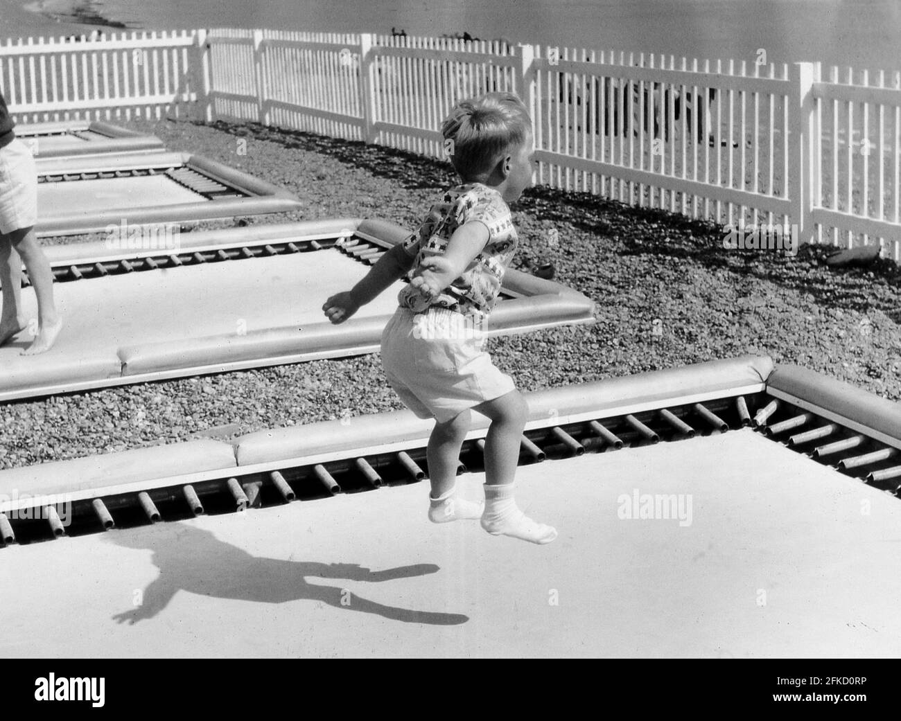 1966, historical, a little boy jumping or bouncing on a trampoline, one of several, in an enclosed, fenced area on a pebbly beach beside the sea. There are many health benefits with using trampolines and bouncing on them is also great fun. Stock Photo