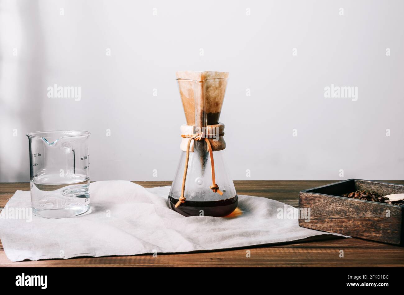 Manual glass coffee pot with filter and coffee on a gray cloth on a wooden table with coffee bean container and glass measuring jug on the sides Stock Photo
