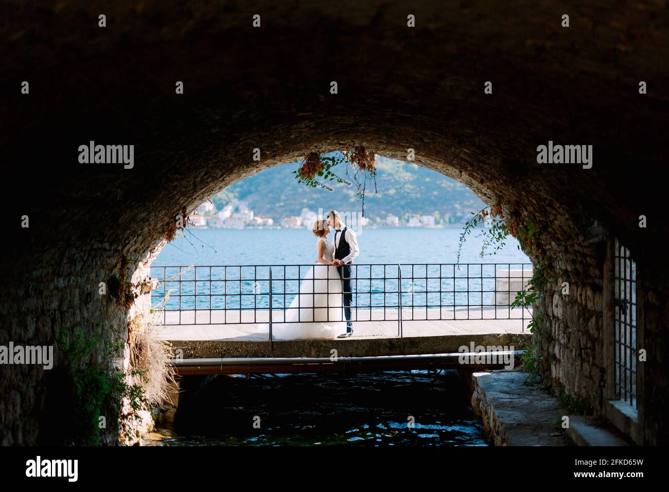The bride and groom hug and kiss on the bridge under the arch, behind ...