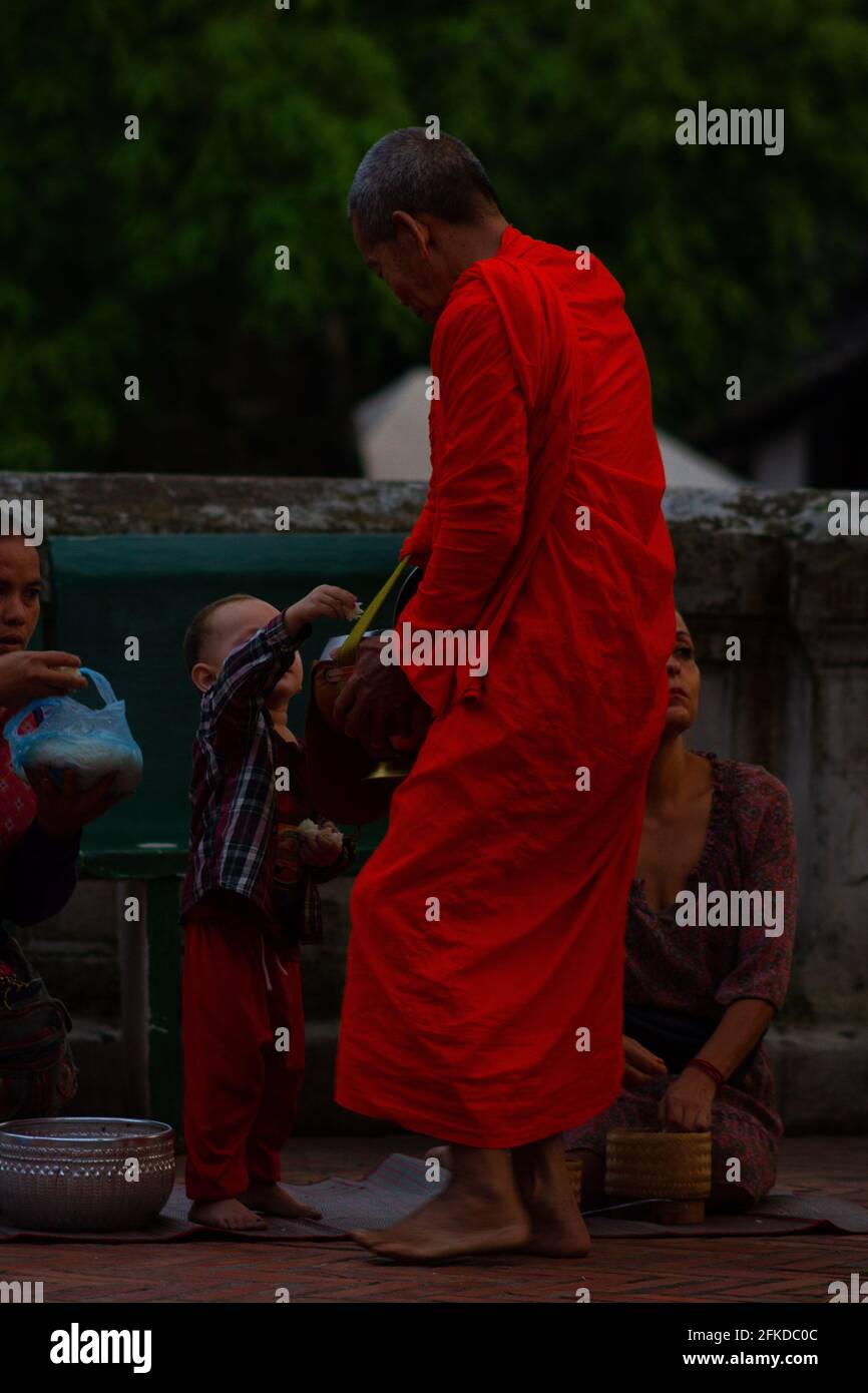 Luang Prabang, Laos - July 6, 2016:  A Buddhist monk accepts rice given to him by a child during the early morning alms giving ceremony of Sai Bat. Stock Photo