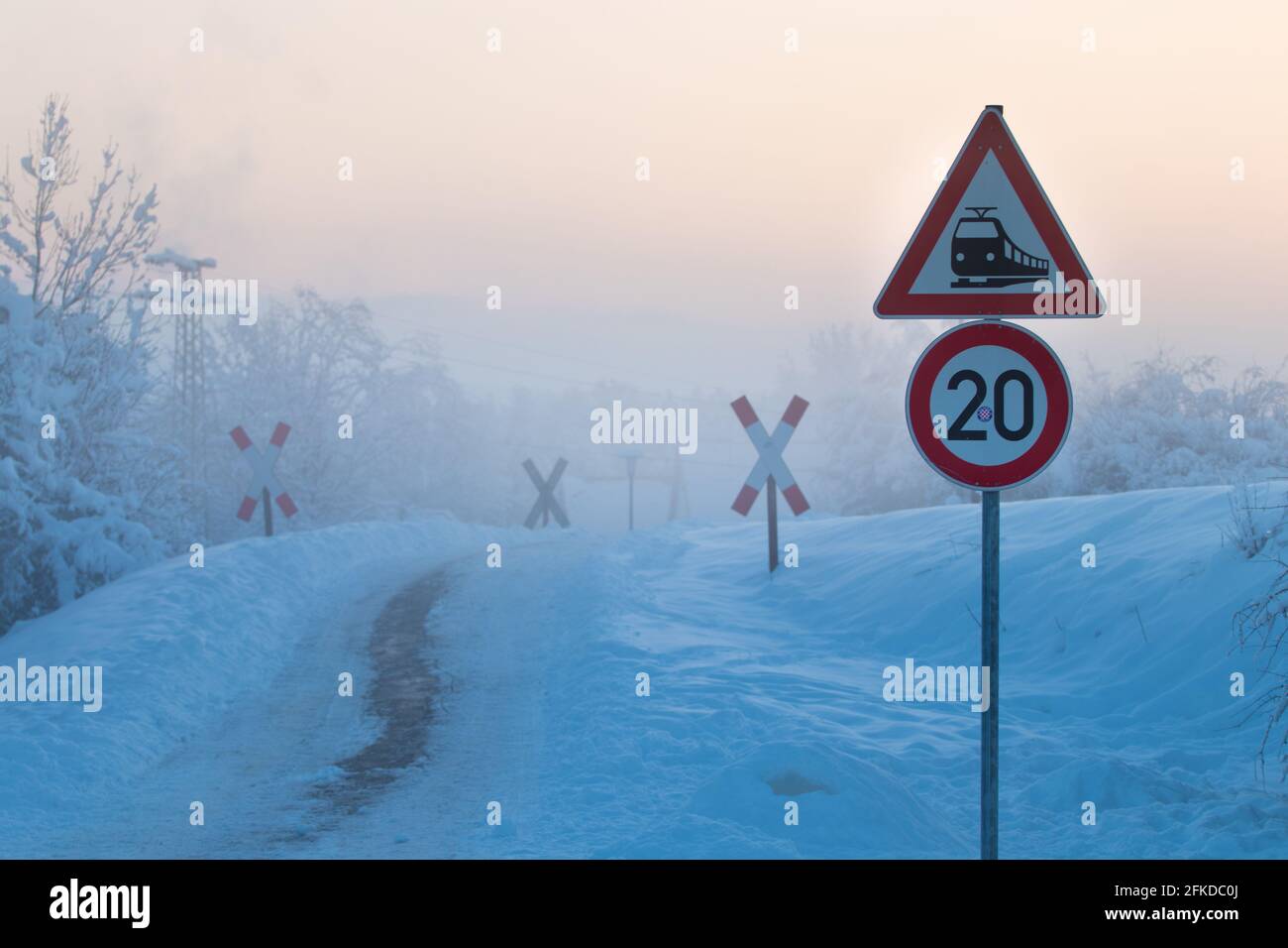 road signs for railway crossing  along a small icy road in  foggy sunset scenery with milky pink sky Stock Photo