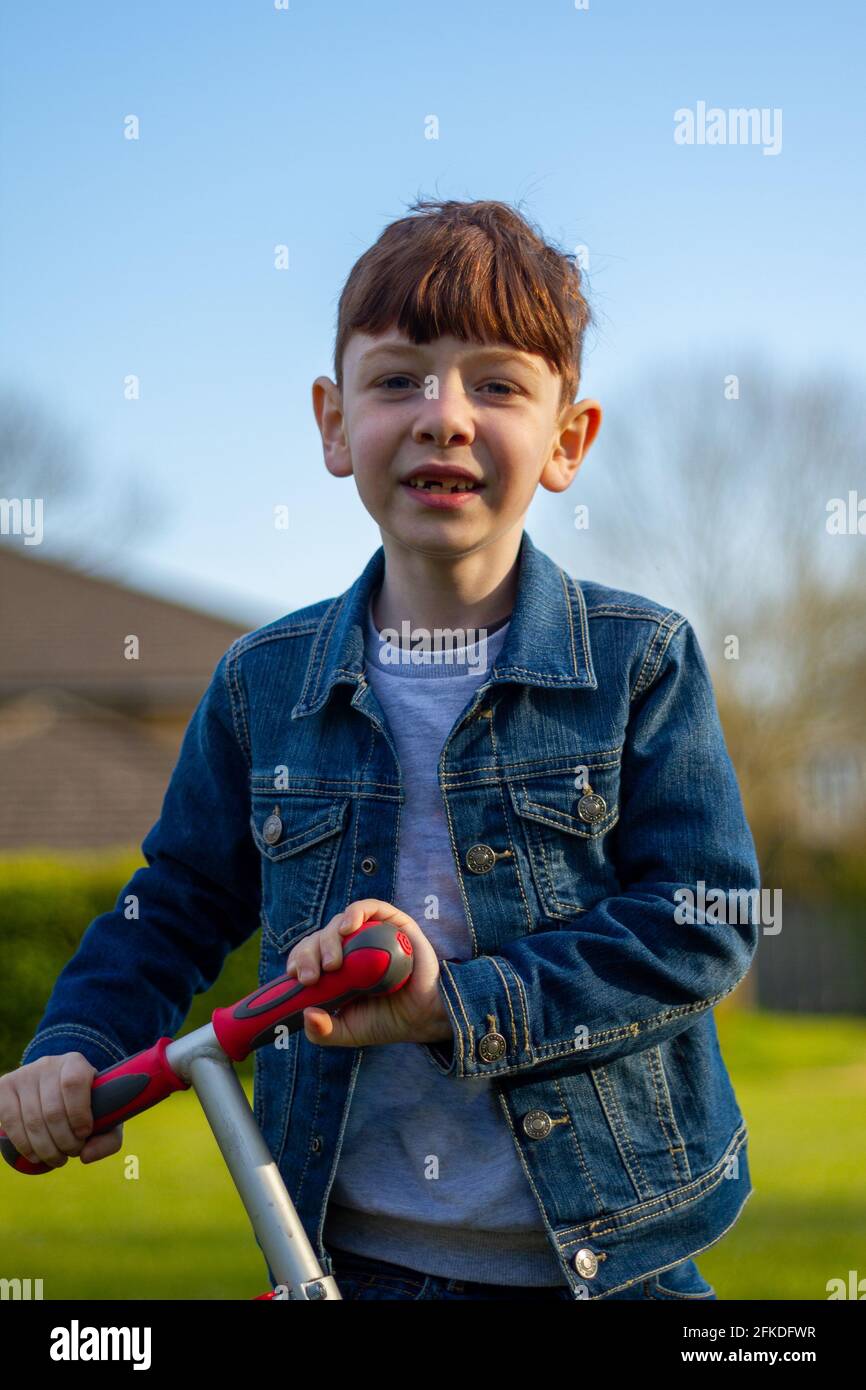 Medium-shot of a cute, redhead boy wearing jeans and a blue denim jacket  playing with a scooter in a lawn on a sunny day Stock Photo - Alamy