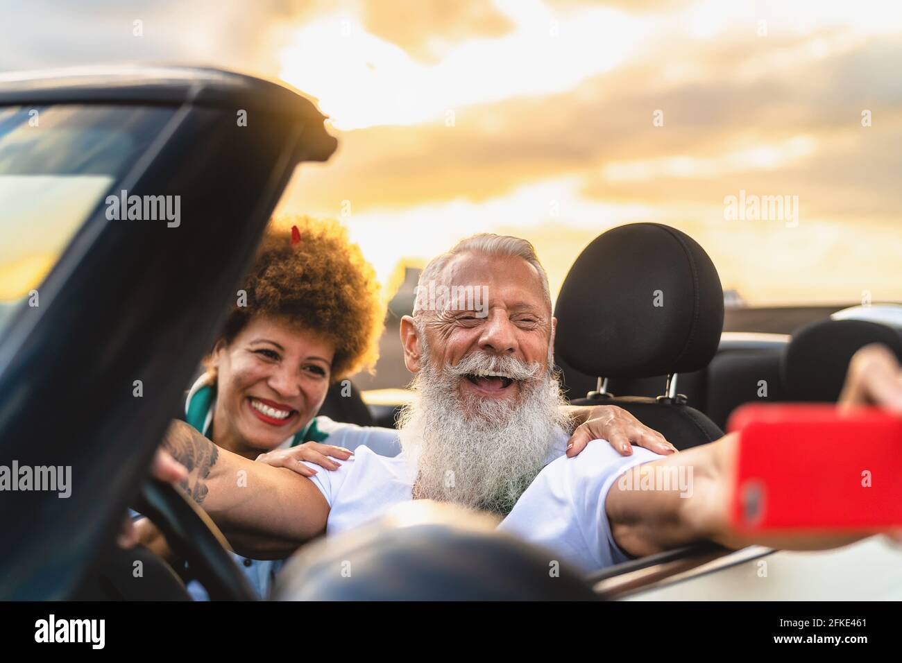 Happy senior couple having fun on new convertible car roadtrip taking selfie with mobile smartphone - Travel people lifestyle concept Stock Photo