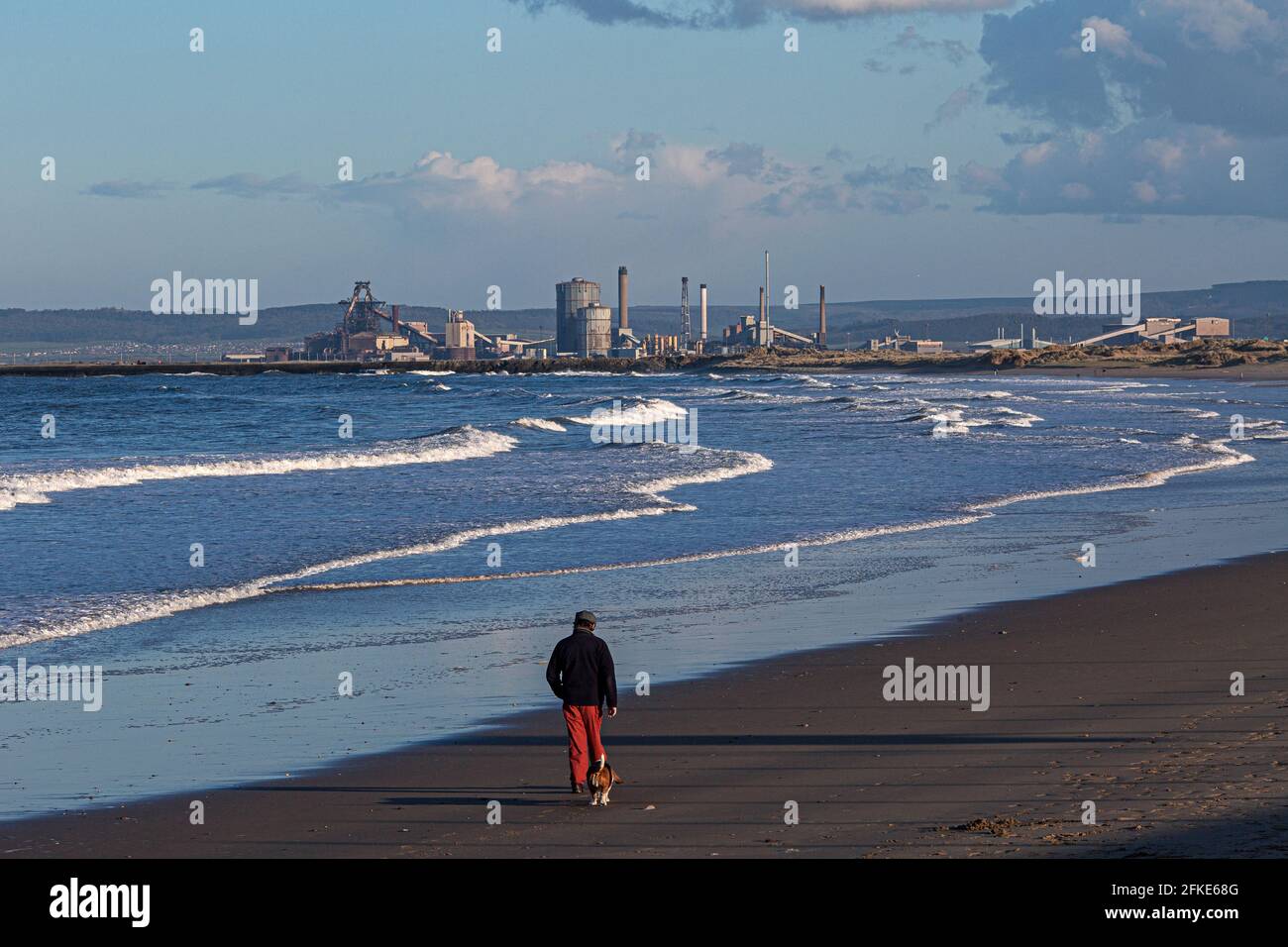 Man and his old dog walking along the beach with the Redcar steelworks in distance , England, UK Stock Photo