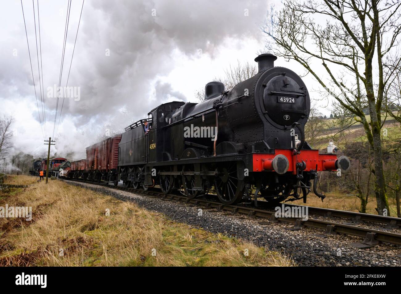 Historic steam freight train or loco puffing smoke clouds (engine driver on footplate, goods wagons, man trackside) - KWVR, Yorkshire, England, UK. Stock Photo