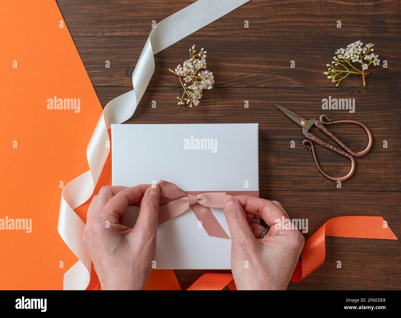 Female hands make a pink bow on a white box placed on a wooden table with flowers and colorful ribbons Stock Photo