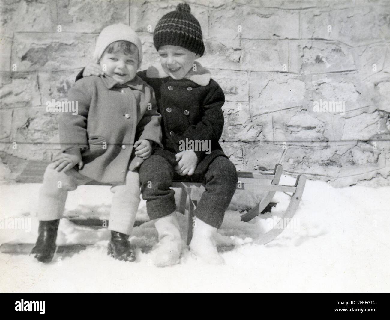 Two siblings sitting on simple sleigh hugging and smiling. Archival photo circa 1969. Stock Photo