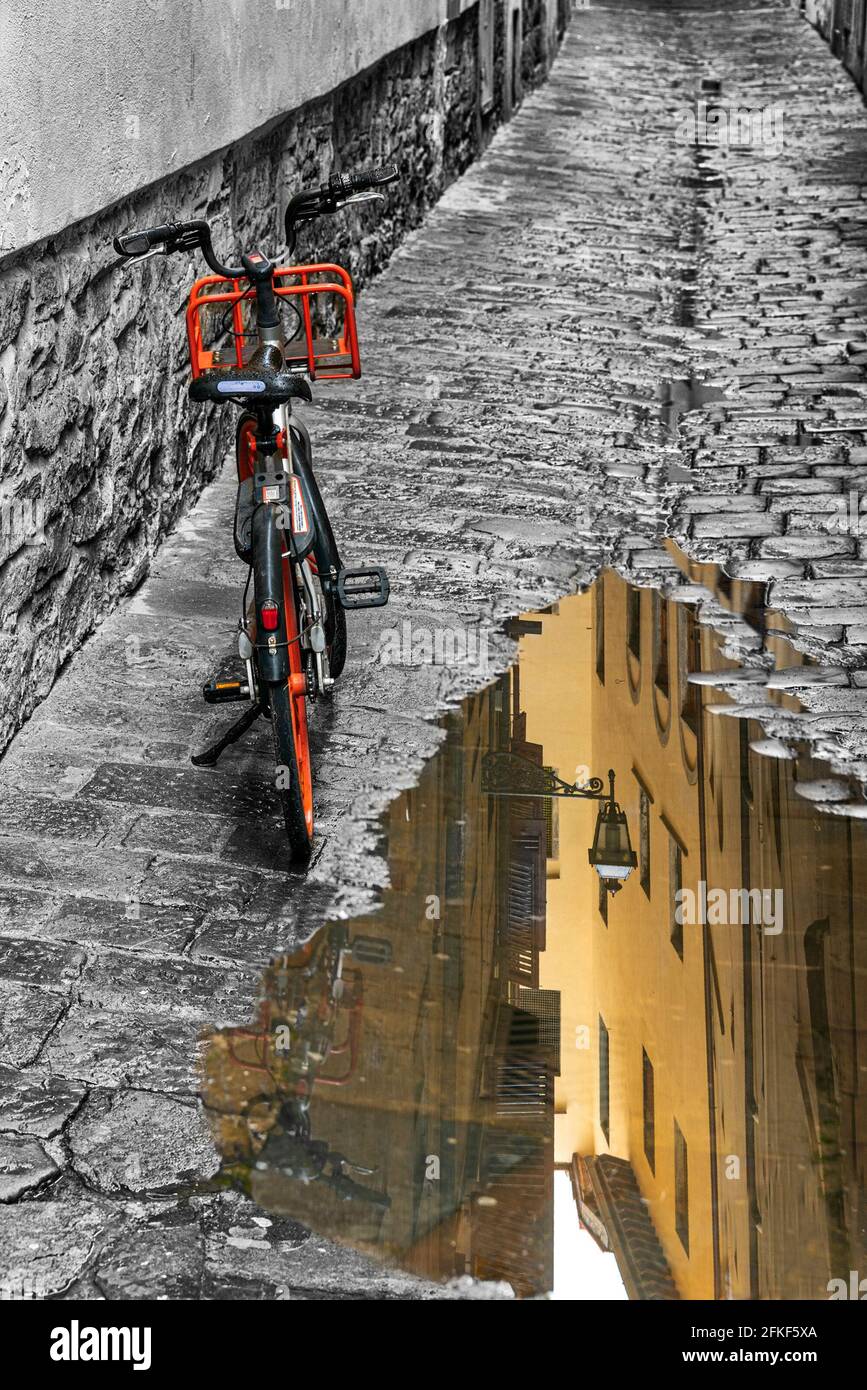 a bike is parked in an alley of Firenze with a puddle reflecting the houses of via delle Bombarde a cross street of Borgo Santi Apostoli Stock Photo