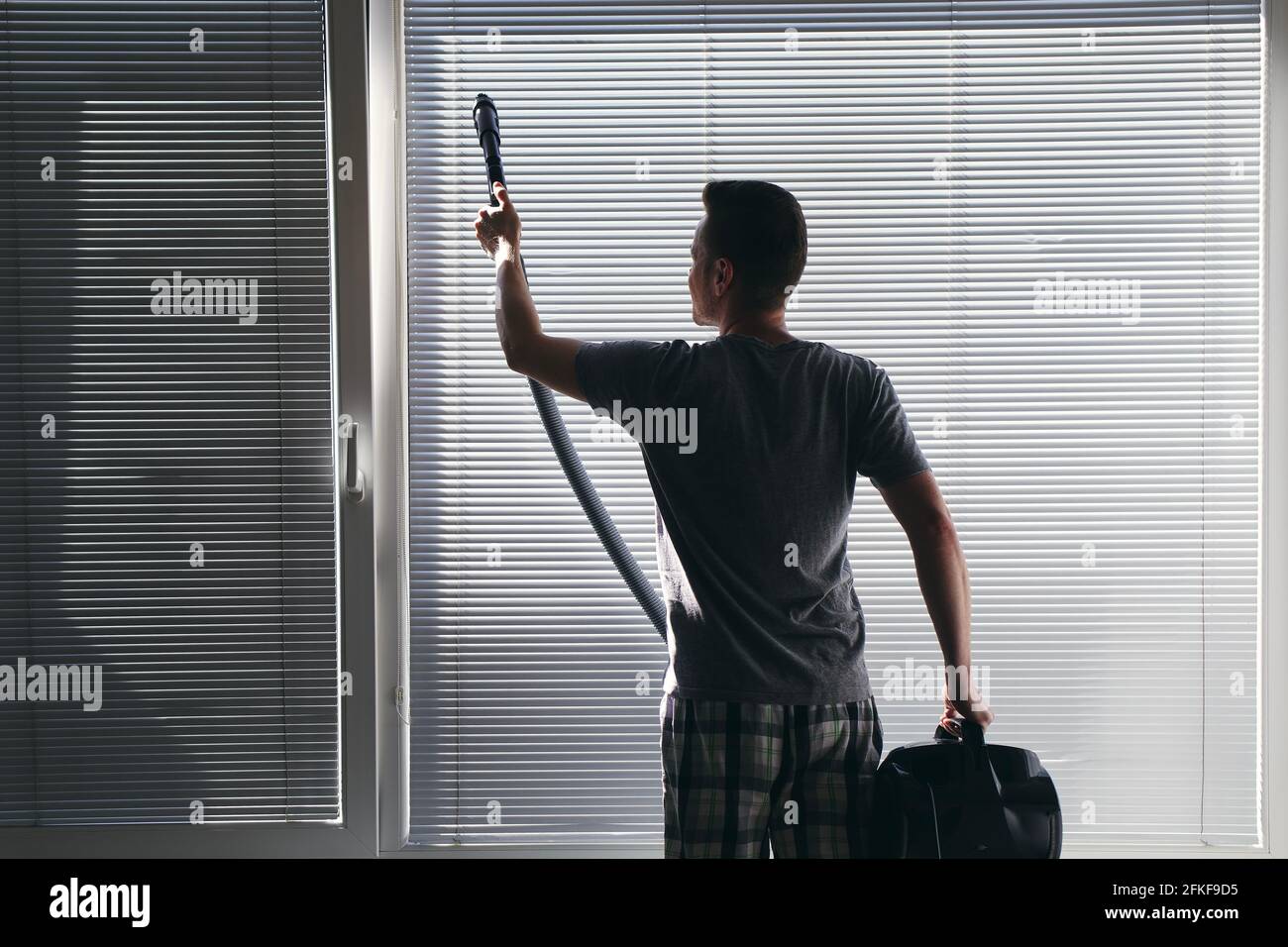 Man cleaning dust from window blind by vacuum cleaner at home. Themes housework and housekeeping. Stock Photo