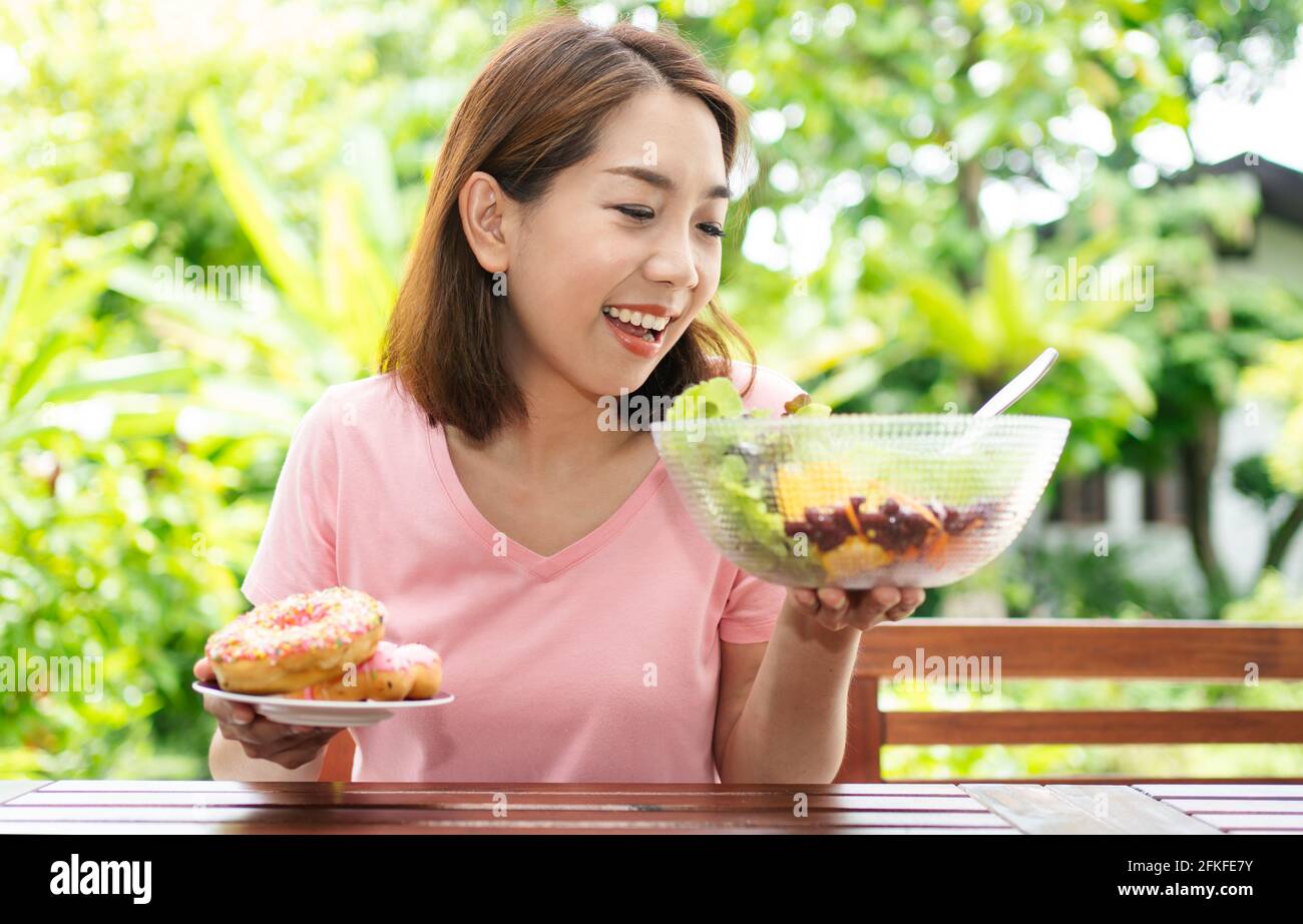 The happy beautiful Asian healthy middle aged woman sitting on the balcony beside the garden and choose between a donut and a vegetable salad. Concept Stock Photo