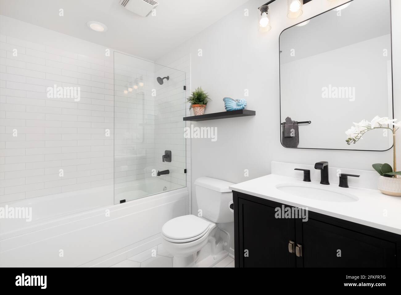 A small modern bathroom with a dark vanity, square mirror,  white subway tiles line the bathtub and shower with black faucets. Stock Photo