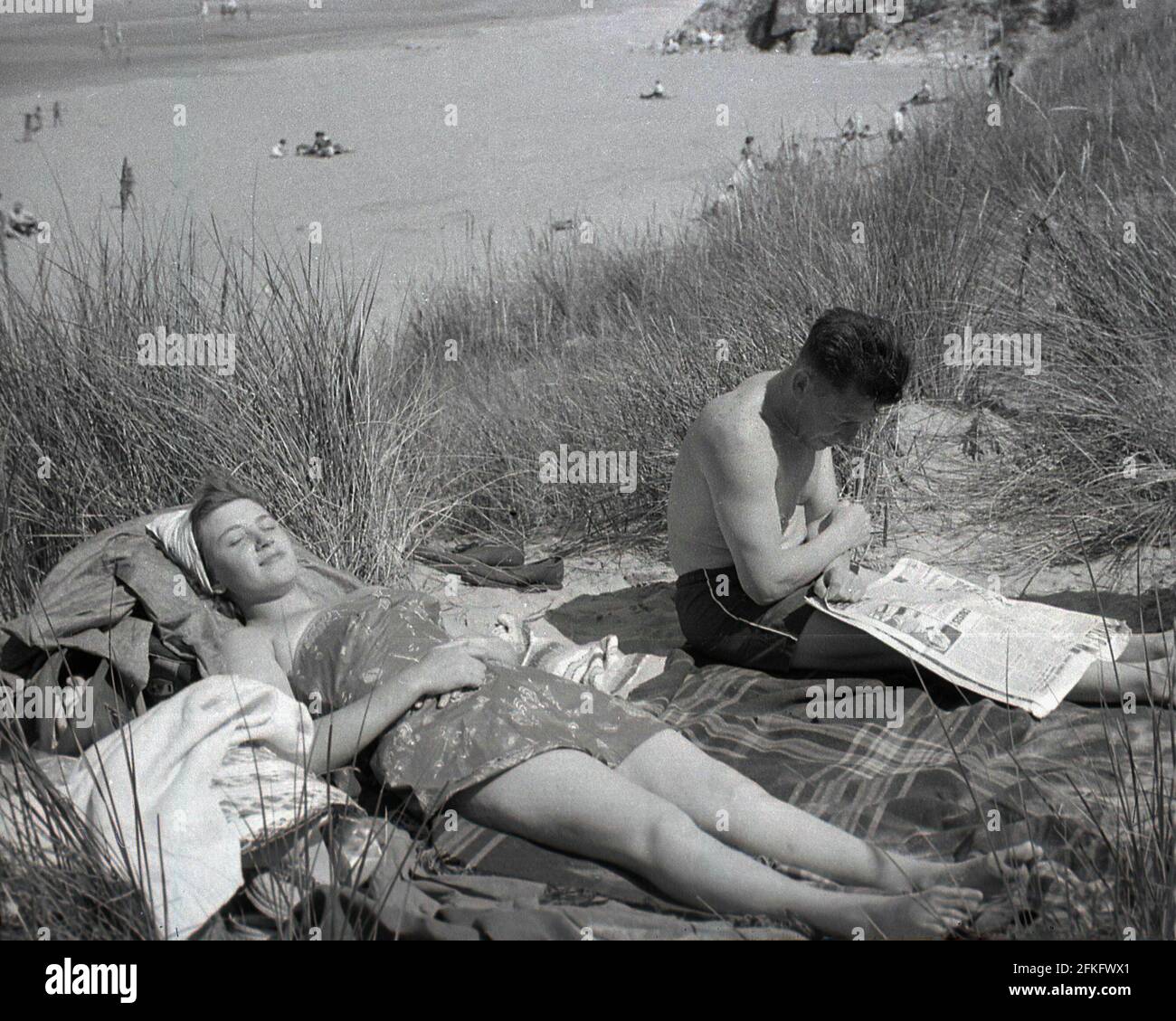 1960s, historical, a couple relaxing on a rug on some sand dunes by a sandy beach, the lady sunbathing, the man sitting reading a broadsheet newspaper, England, UK. Stock Photo