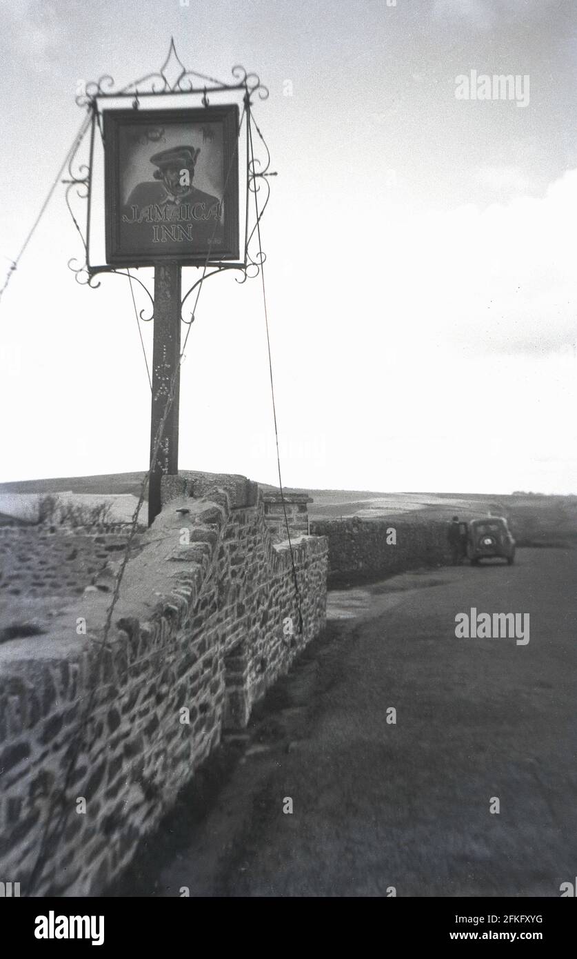 1950s, In a remote lcoation on Bodmin Moor, beside a stone wall and narrow road, a sign for the Jamacia Inn tavern, Bolventor, Cornwall, England, UK. Dating back to 1750, this old coaching inn on Bodmin Moor became famous when featured in the 1936 novel 'Jamaica Inn' by author Daphne du Maurier. Remote and isolated, it was originally built as a coaching inn for travellers using the turnpike road between Launceston and Bodmin over the wild Moor and later became known as a place where contraband - rum, brandy, tea in those days - were smuggled in from aboard, onto the Cornish coast. Stock Photo