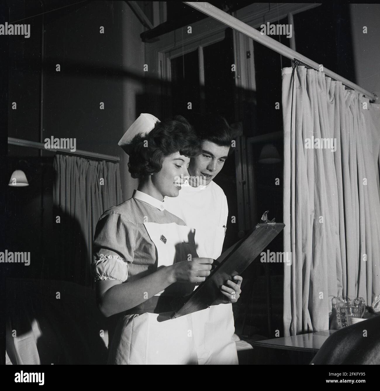 1960s, historical, doing the rounds......late evening and on a hospital ward, an attractive female nurse holding a wooden clipboard, smiles as she shows a young male doctor, a patient's chart, England, UK. Ward visits are an important part of a doctor's routine in assessing patients needs and the support of nursing staff in this role is vital for good and effective communication. Stock Photo