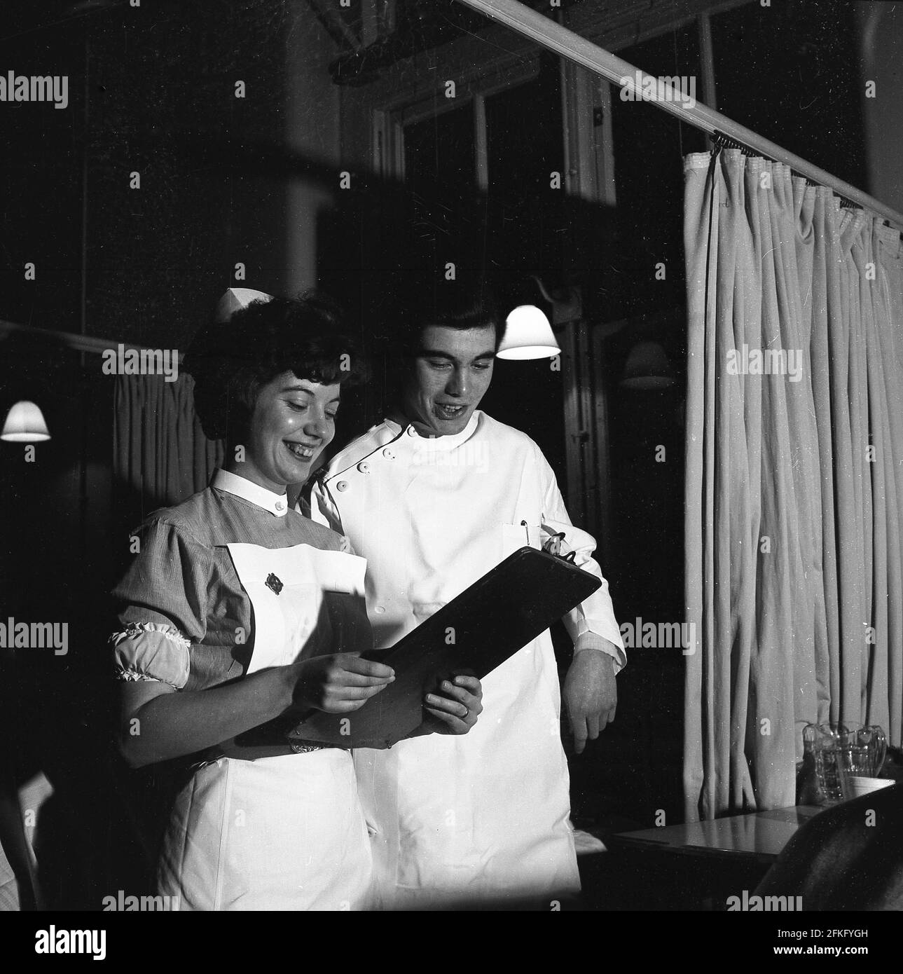 1960s, historical, doing the rounds......late evening and on a hospital ward, an attractive female nurse holding a wooden clipboard, smiles as she shows a young male doctor, a patient's chart, England, UK. Nighttime ward rounds are a part of a doctor's routine in assessing care for patients and the support of nursing staff in this role is vital for good and effective communication. Stock Photo