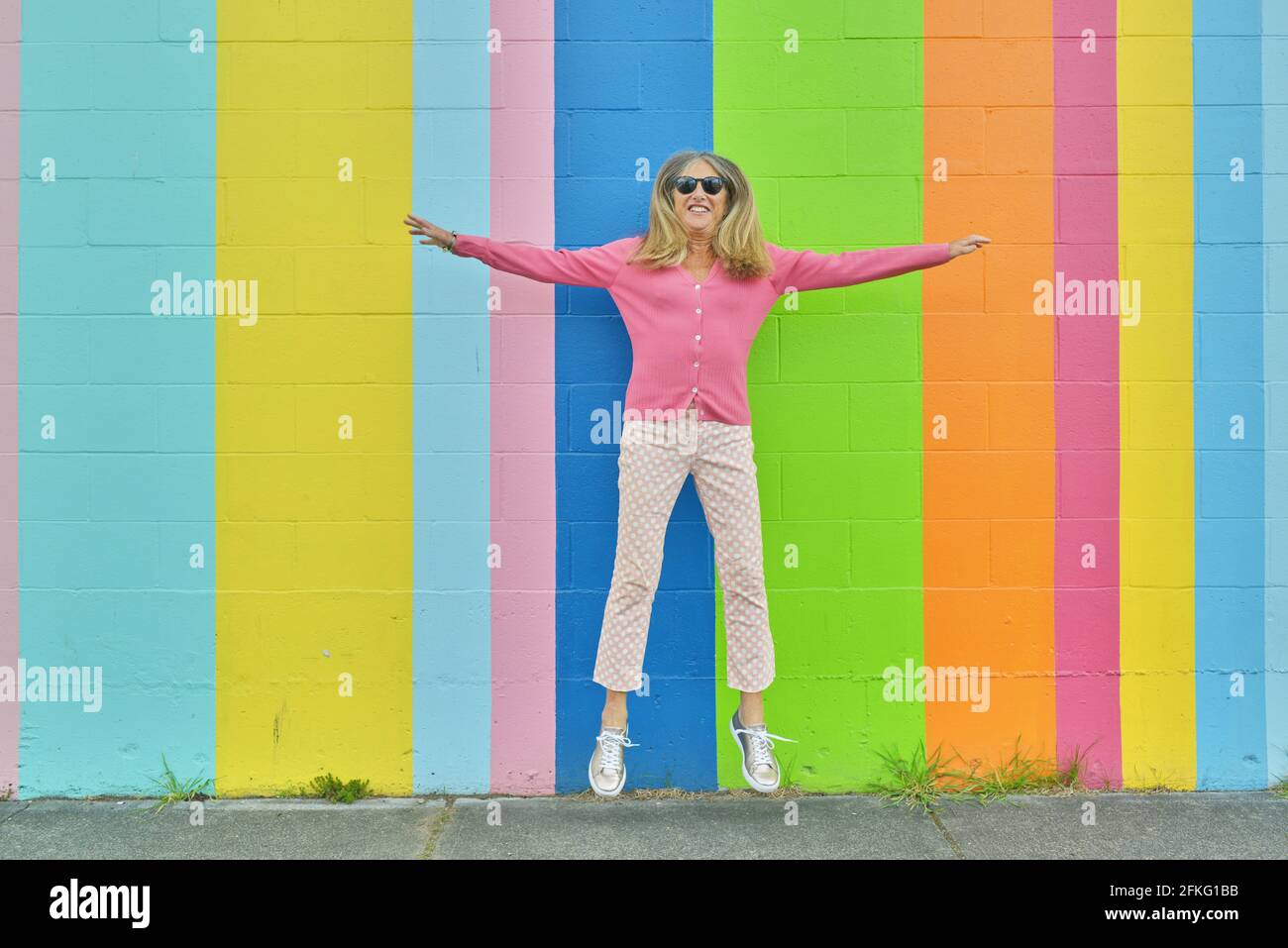 Attractive senior woman jumping  against brightly colored striped wall outdoors Stock Photo