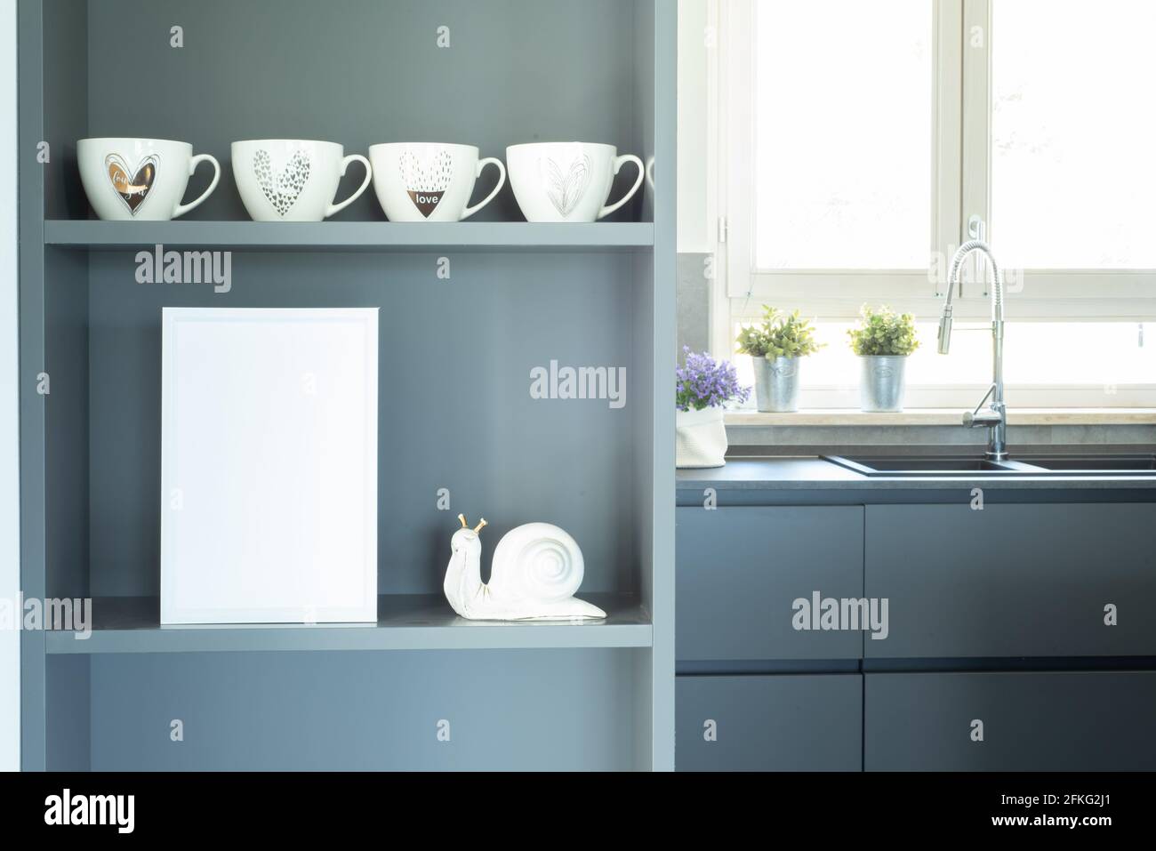 Elegant and cozy gray kitchen. Blank white frame mockup on the furniture shelf in the foreground. Modern interior illuminated by natural light from th Stock Photo