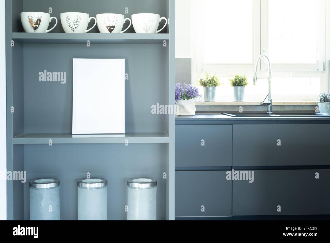 Elegant and cozy gray kitchen. Blank white frame mockup on the furniture shelf in the foreground. Modern interior illuminated by natural light from th Stock Photo
