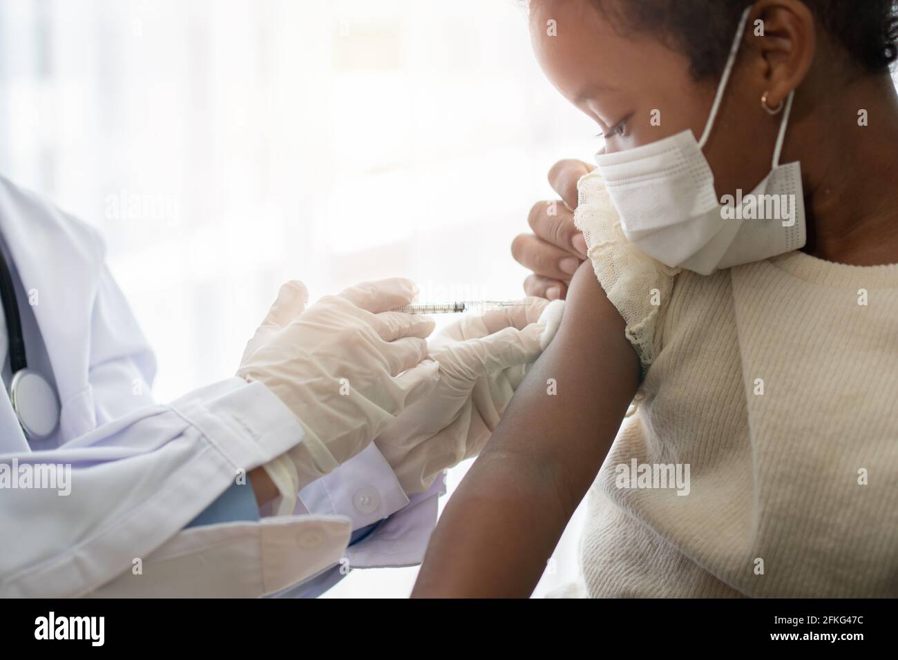 Asian doctor user syringe needle for injection vaccinate,  African American children are being covid vaccinated for prevention coronavirus and flu. Co Stock Photo