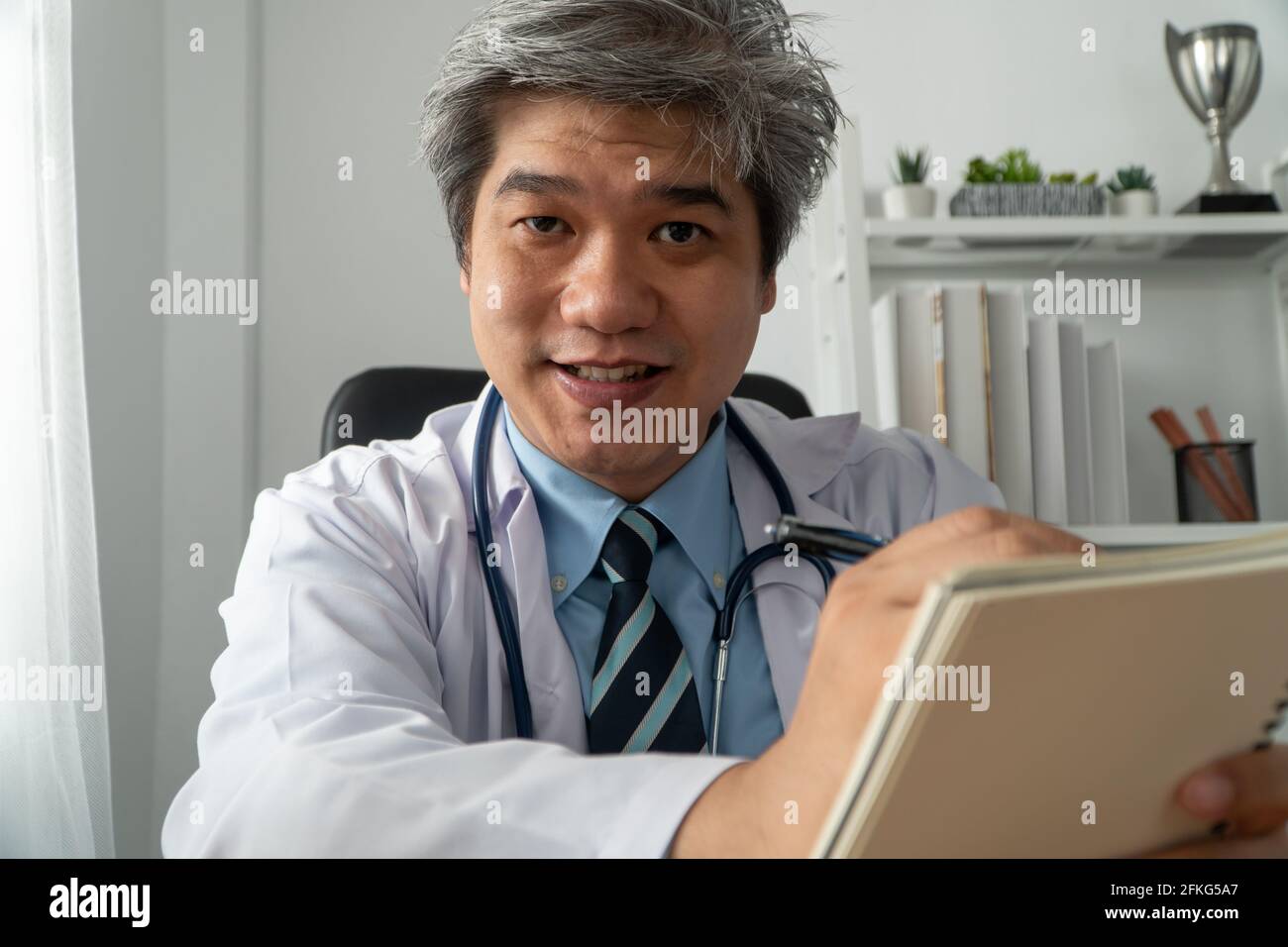 Asian doctor is online visiting with a patient on the internet application and make a note of the symptoms and explain how to treat the initial diseas Stock Photo