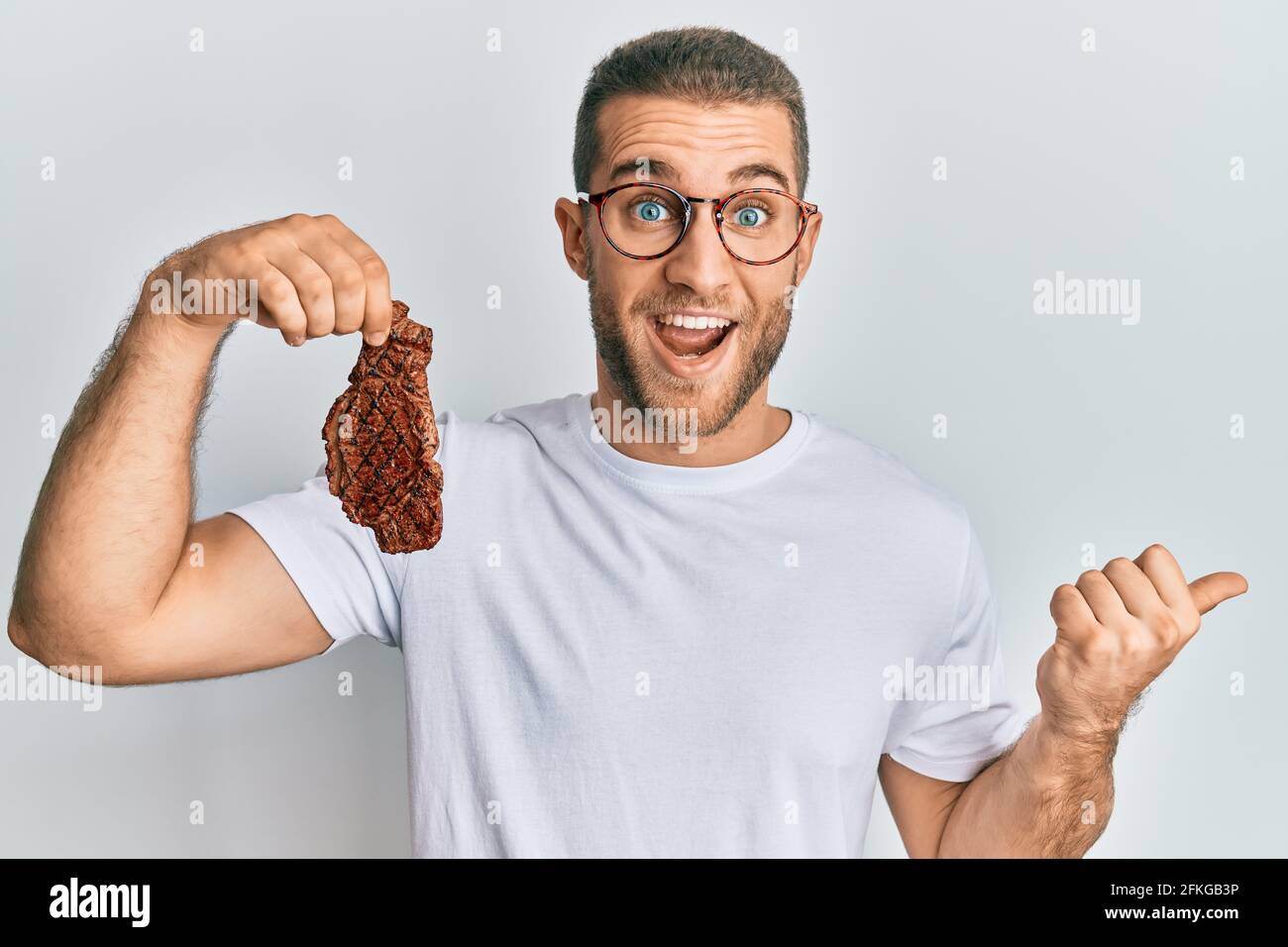 Young caucasian man holding beef steak pointing thumb up to the side ...