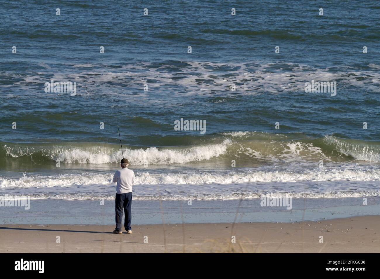 Older white man stands alone on a beach and fishes in the ocean on a ...