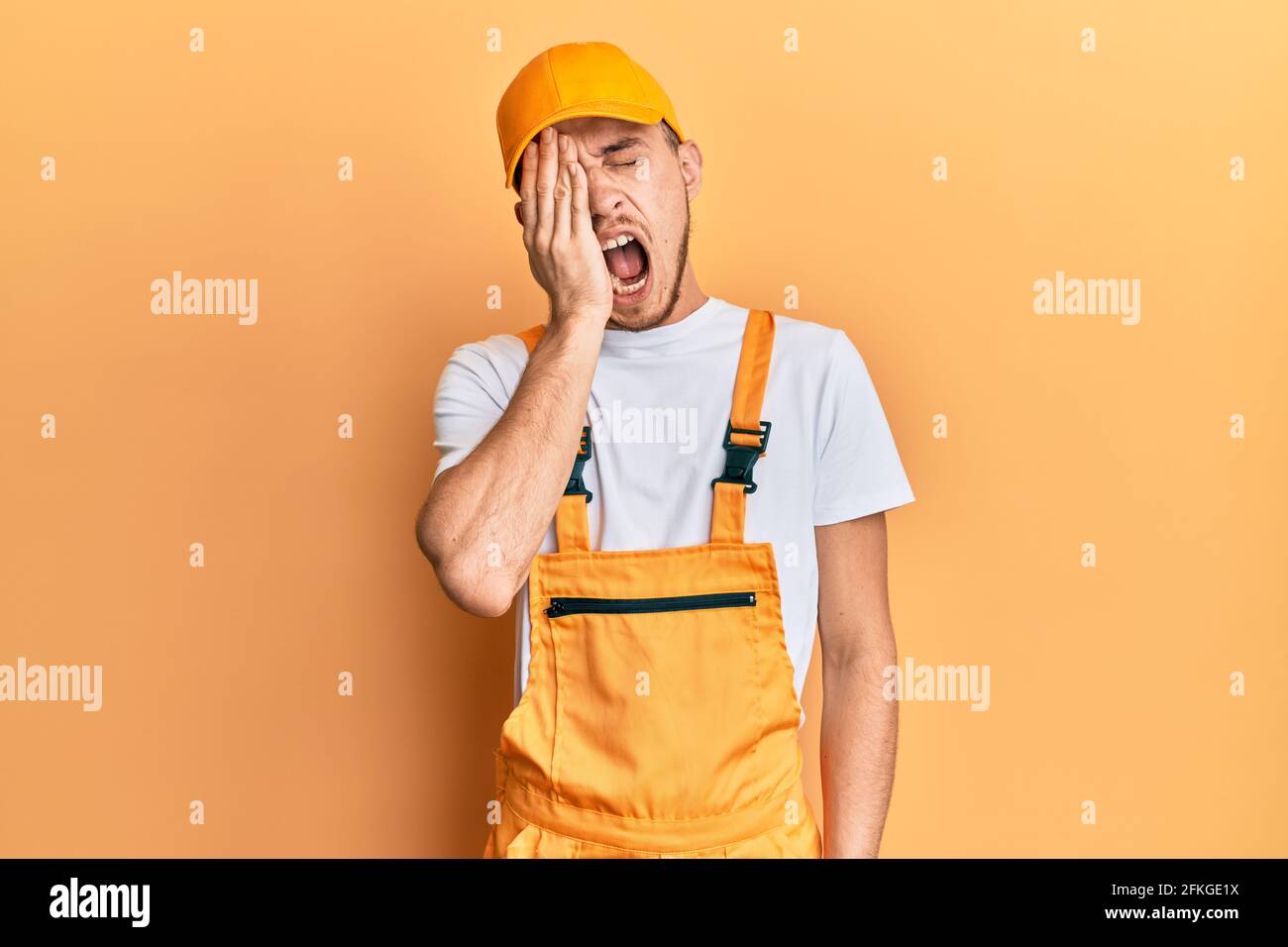 Hispanic young man wearing handyman uniform yawning tired covering half ...