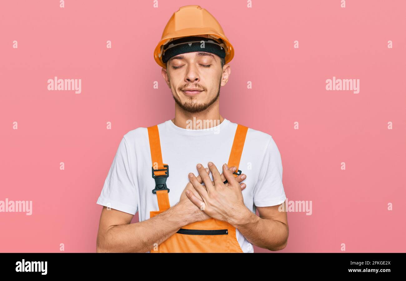 Hispanic young man wearing handyman uniform and safety hardhat smiling ...